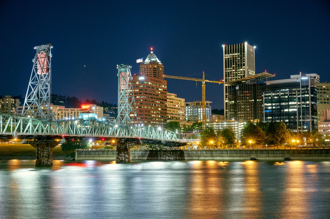 Downtown Portland at night seen from across the Willamette River, with the lit Hawthorne Bridge in the foreground and office towers behind it.