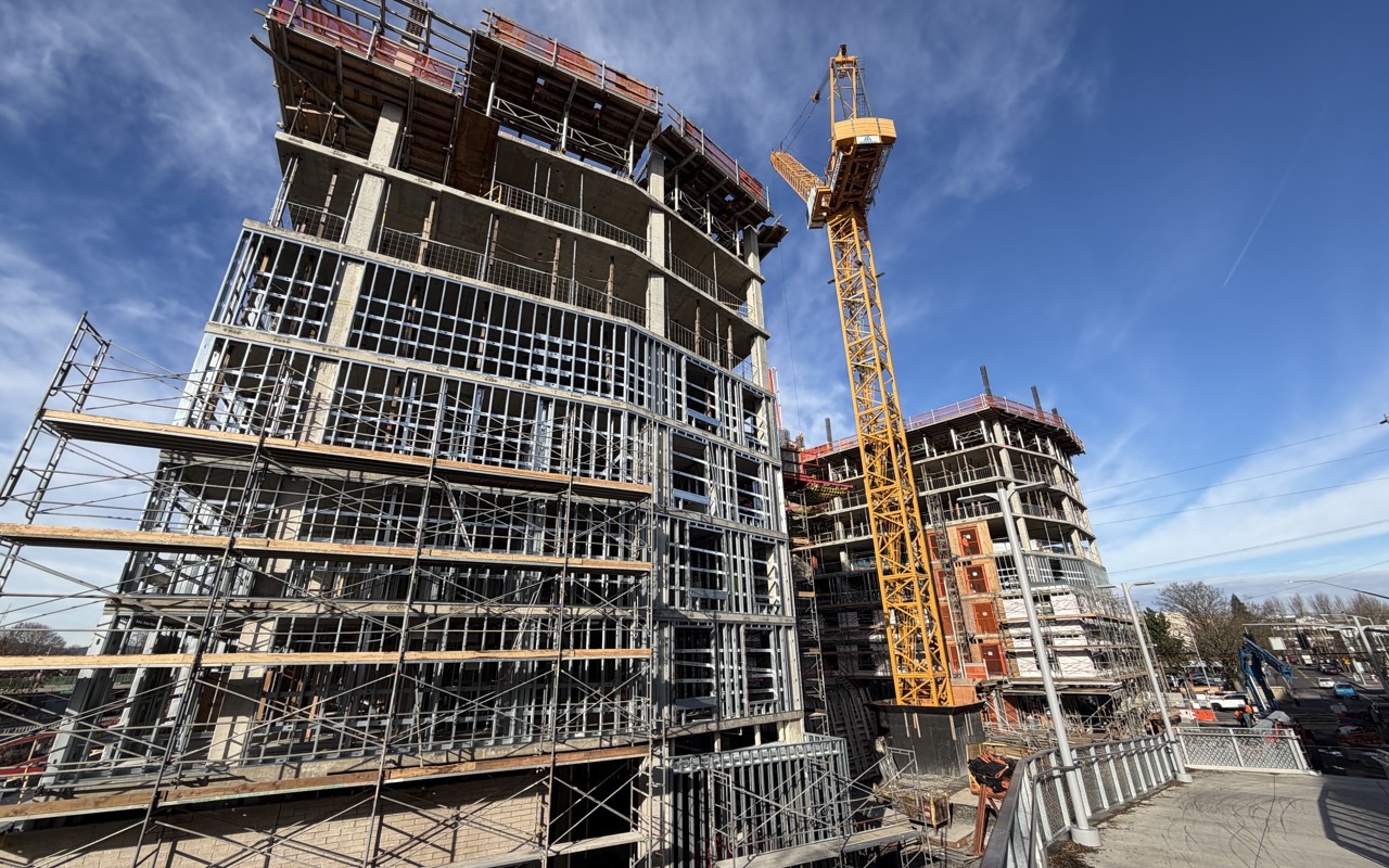 A 12-story apartment building under construction, wrapped in scaffolding and steel framing, with a yellow tower crane alongside.