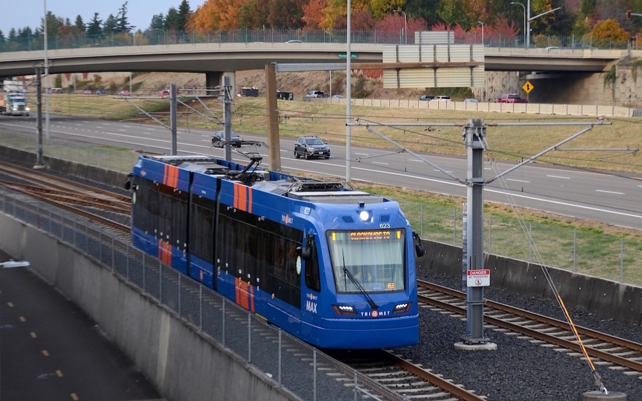 A blue TriMet MAX light rail car running beside the I-205 freeway, its destination sign reading Clackamas TC.